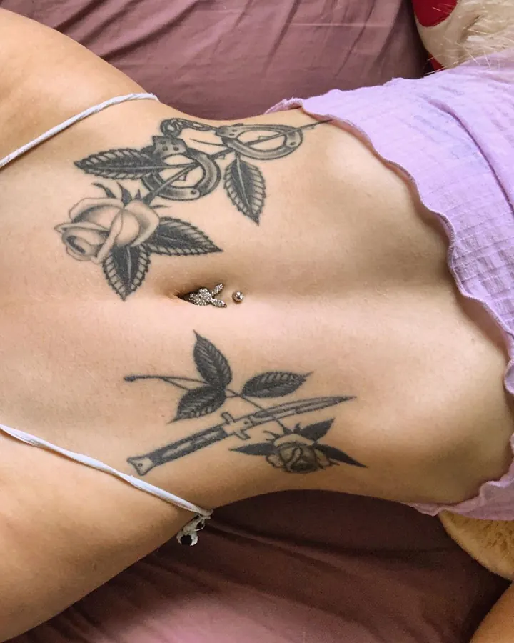 Ventre d'une femme avec un tatouage de rose et de couteau, un piercing au nombril, sous une robe rose