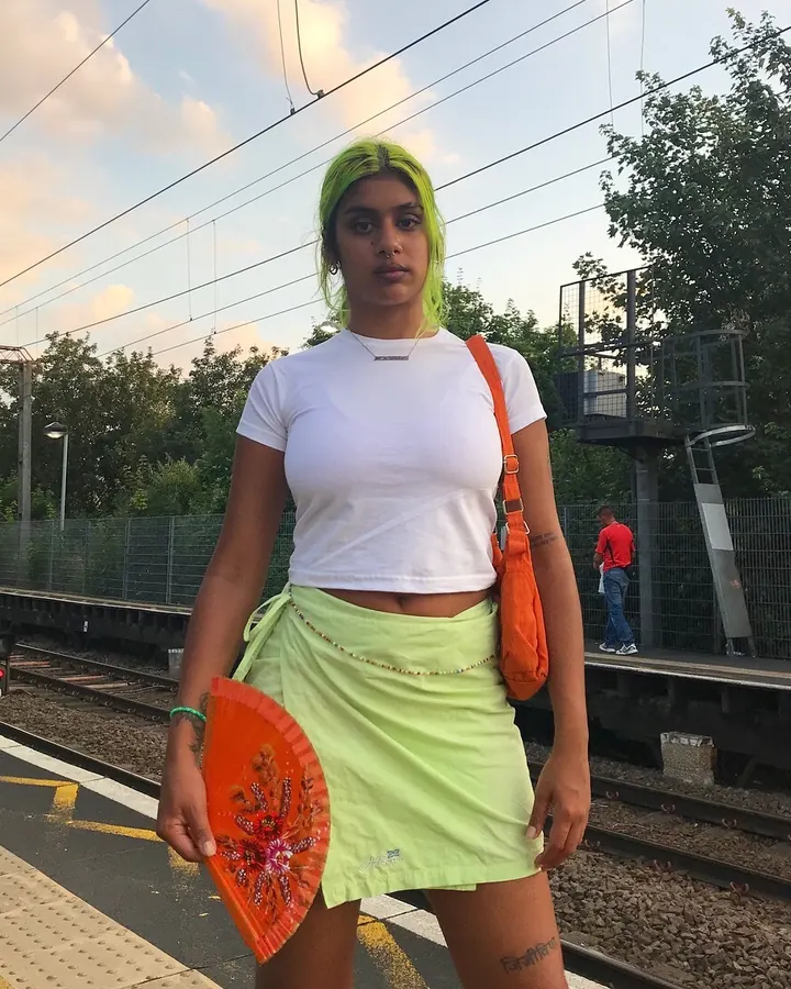 Femme en tenue colorée, debout sur un quai de train, tenant un éventail orange.