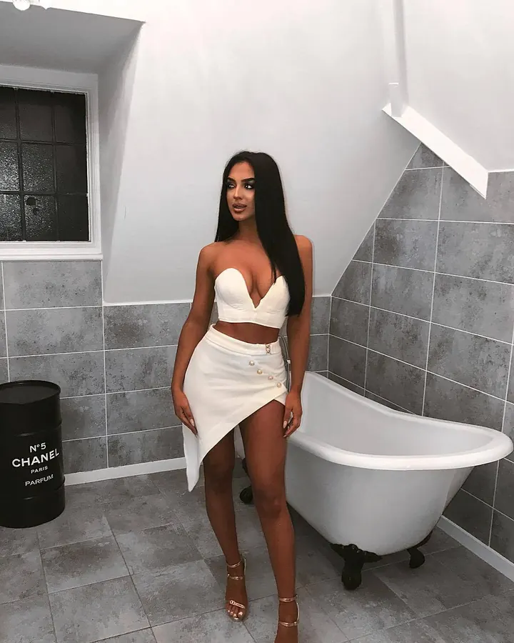 Femme en tenue blanche, bustier et jupe asymétrique, debout près d'une baignoire blanche dans une salle de bains grise.