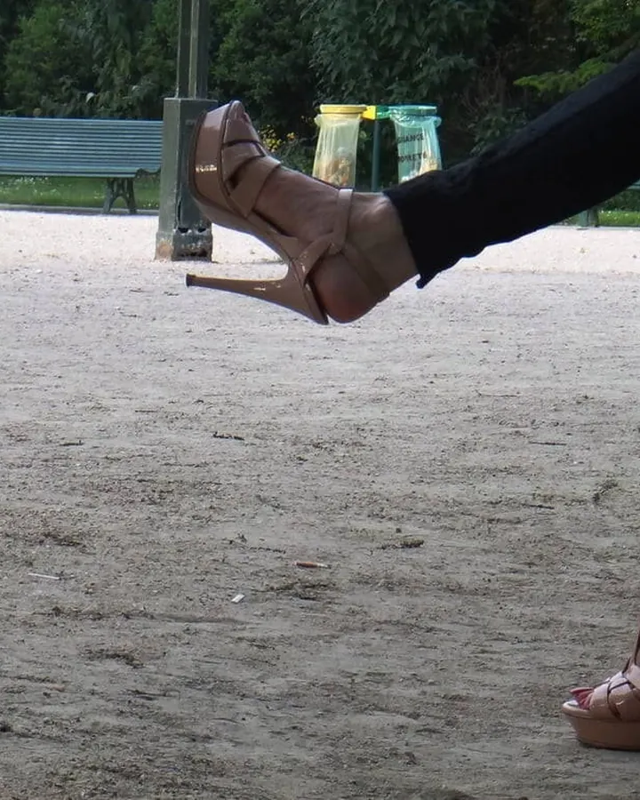 Femme en sandales à talons hauts, pieds sur un sol sablonneux, dans un parc avec bancs en arrière-plan.