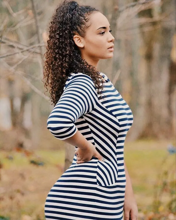 Femme en robe rayée bleu marine et blanche, de profil, les cheveux bouclés en chignon, main sur la hanche.