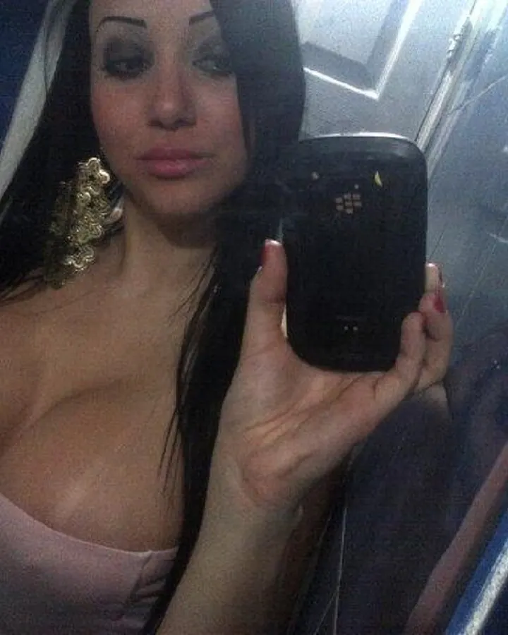 Femme en tenue rose pâle, prenant un selfie dans un miroir, avec un téléphone noir, dans une salle de bain.