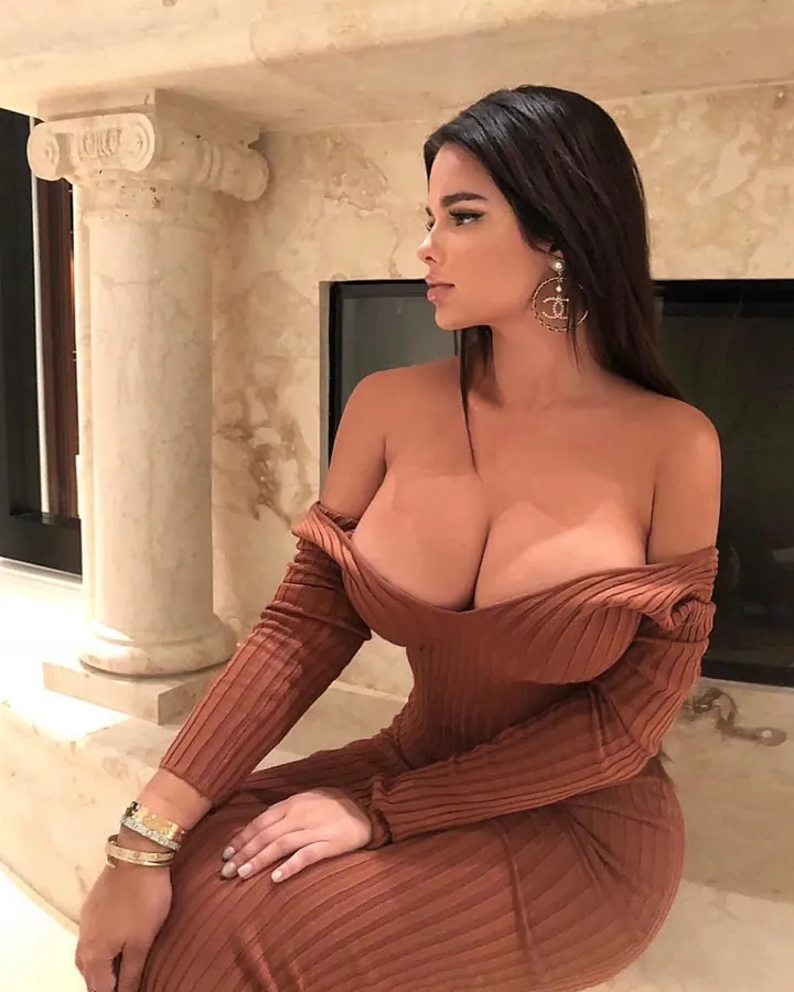 Femme assise sur un rebord en marbre, portant une robe marron moulante, avec des bijoux Chanel, regardant de côté.