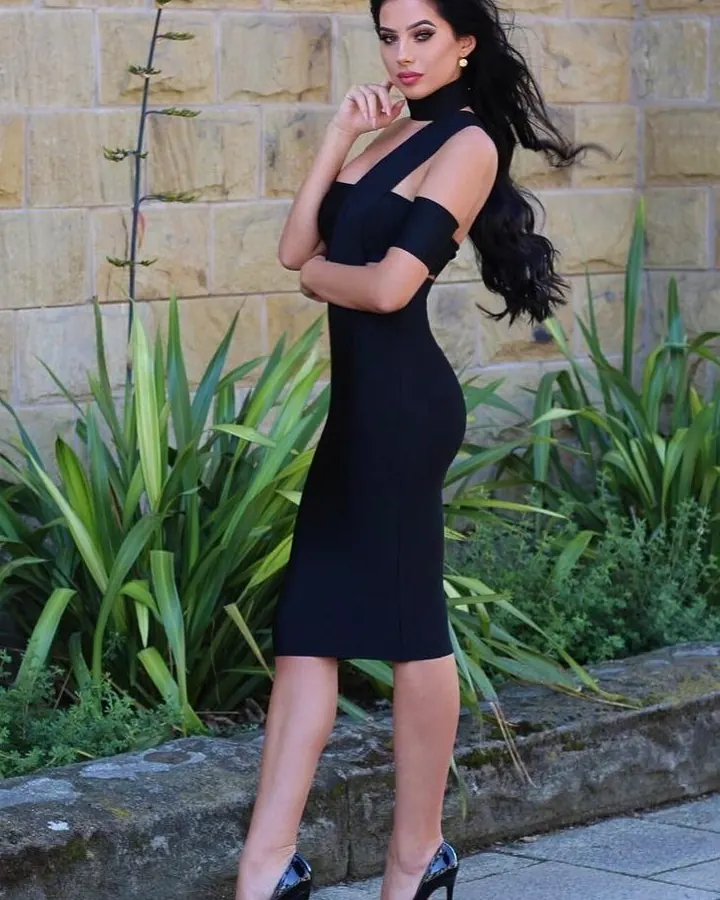 Femme arabe de 35-45 ans, cheveux bruns-noirs, portant une robe noire ajustée, posant près d'un mur en pierre.
