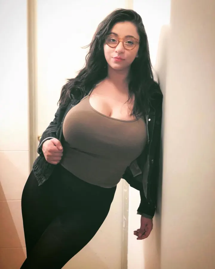 Femme aux cheveux bruns-noirs portant des lunettes et une veste noire, appuyée contre un mur, en haut gris et leggings noirs.